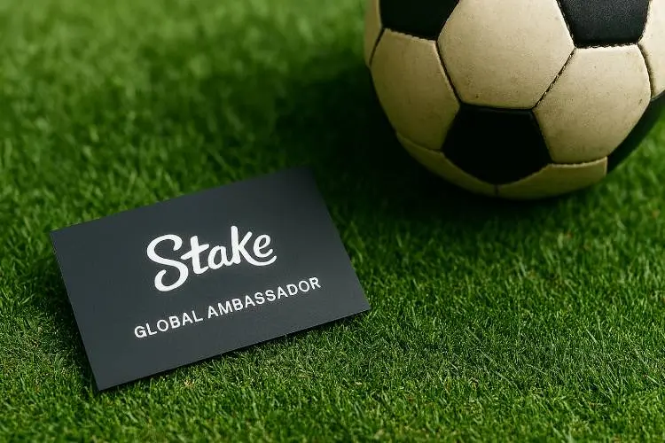 A business card with the Stake logo and “Global Ambassador” title lying on green football turf beside a vintage soccer ball.