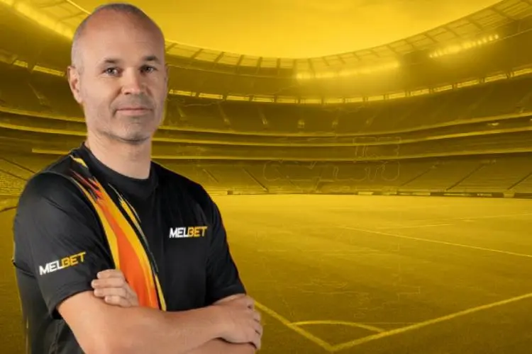 Andrés Iniesta wearing a MelBet jersey stands in a golden-lit football stadium, representing his new role as MelBet brand ambassador.